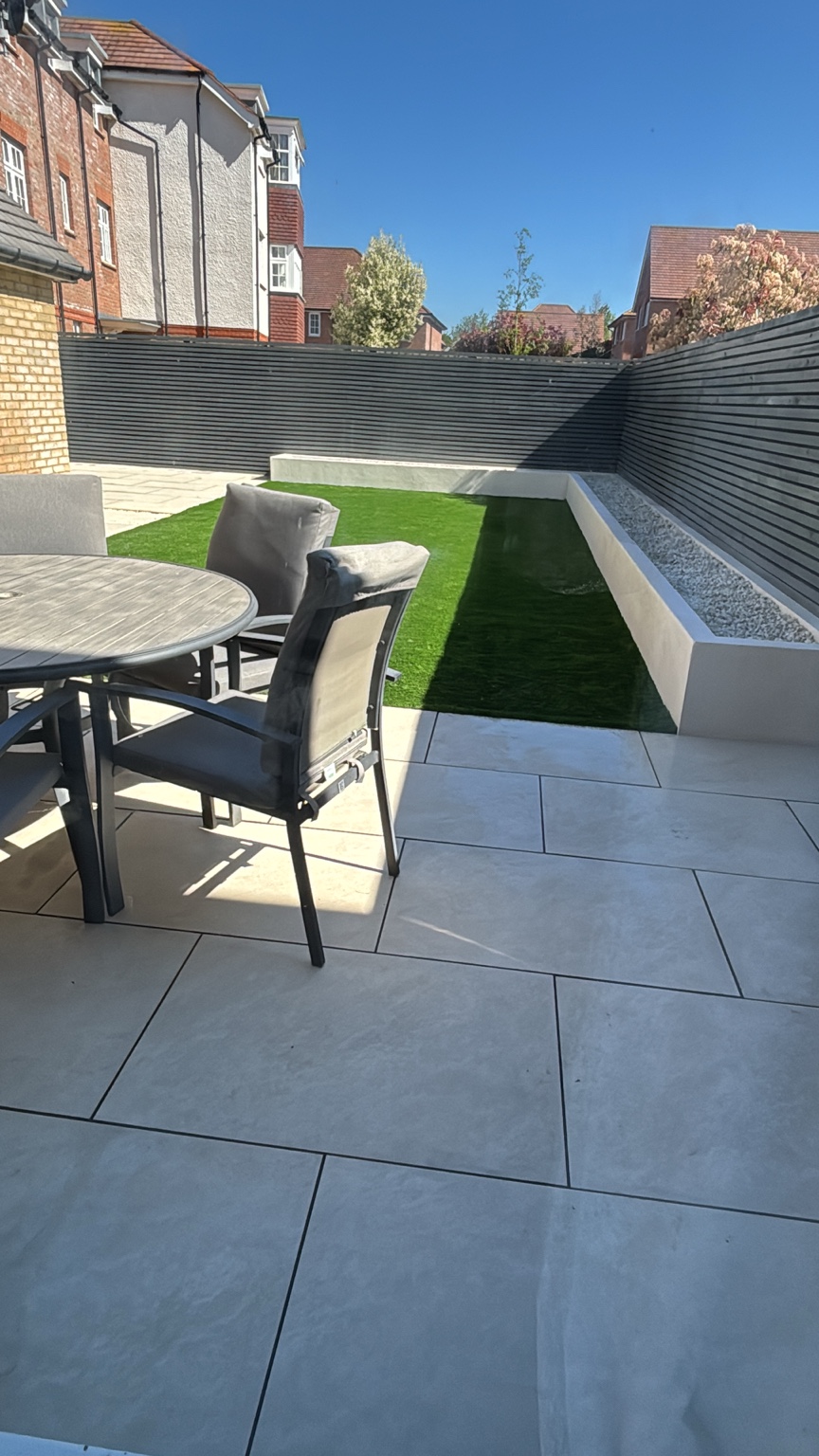 After landscaping: light grey porcelain patio, central artificial grass, a light raised bed and dark slatted fencing